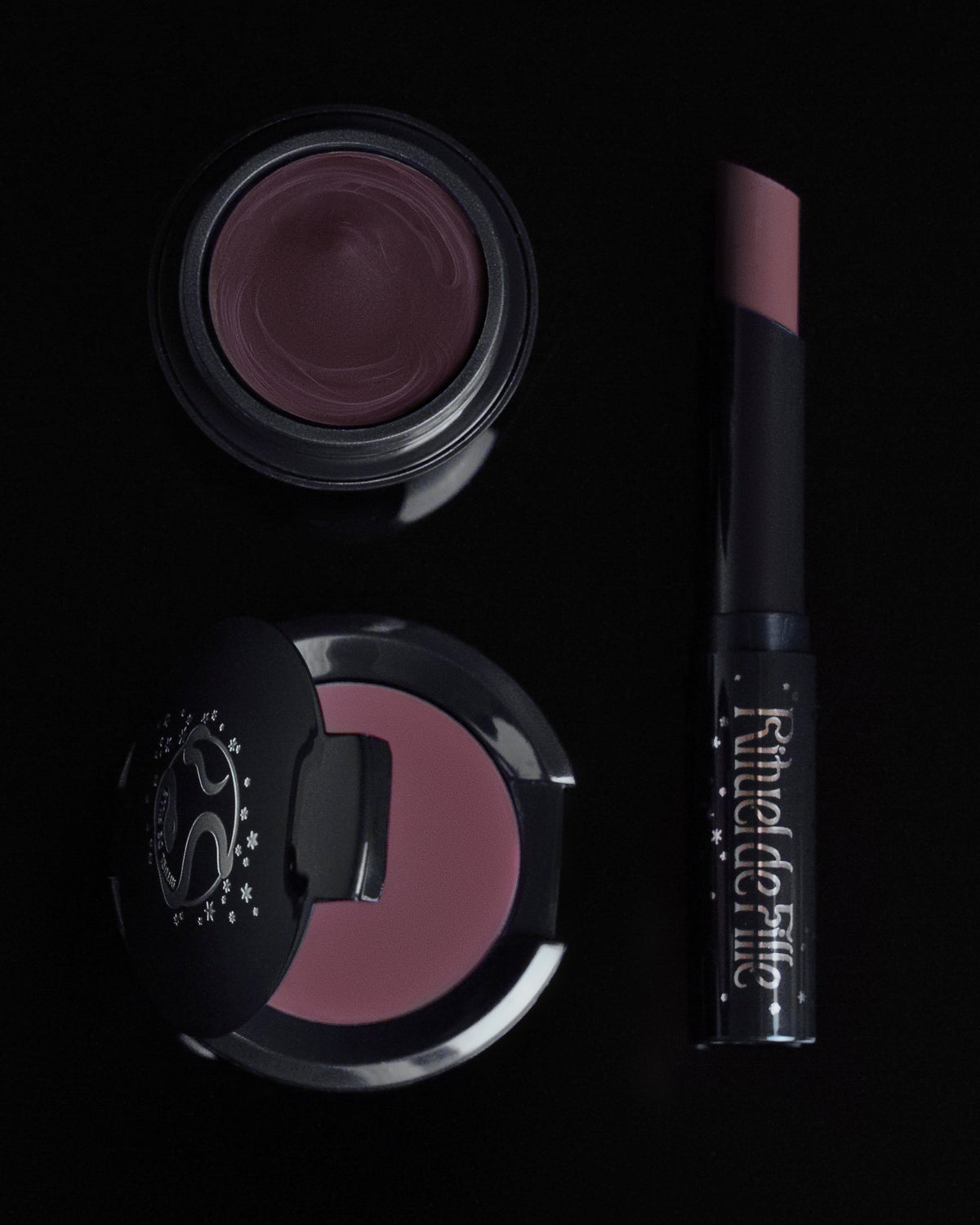 A makeup set featuring a pot of mauve-purple cream product, a matching compact, and a lip product tube labeled 'Rituel de Fille' in a dark studio setting.