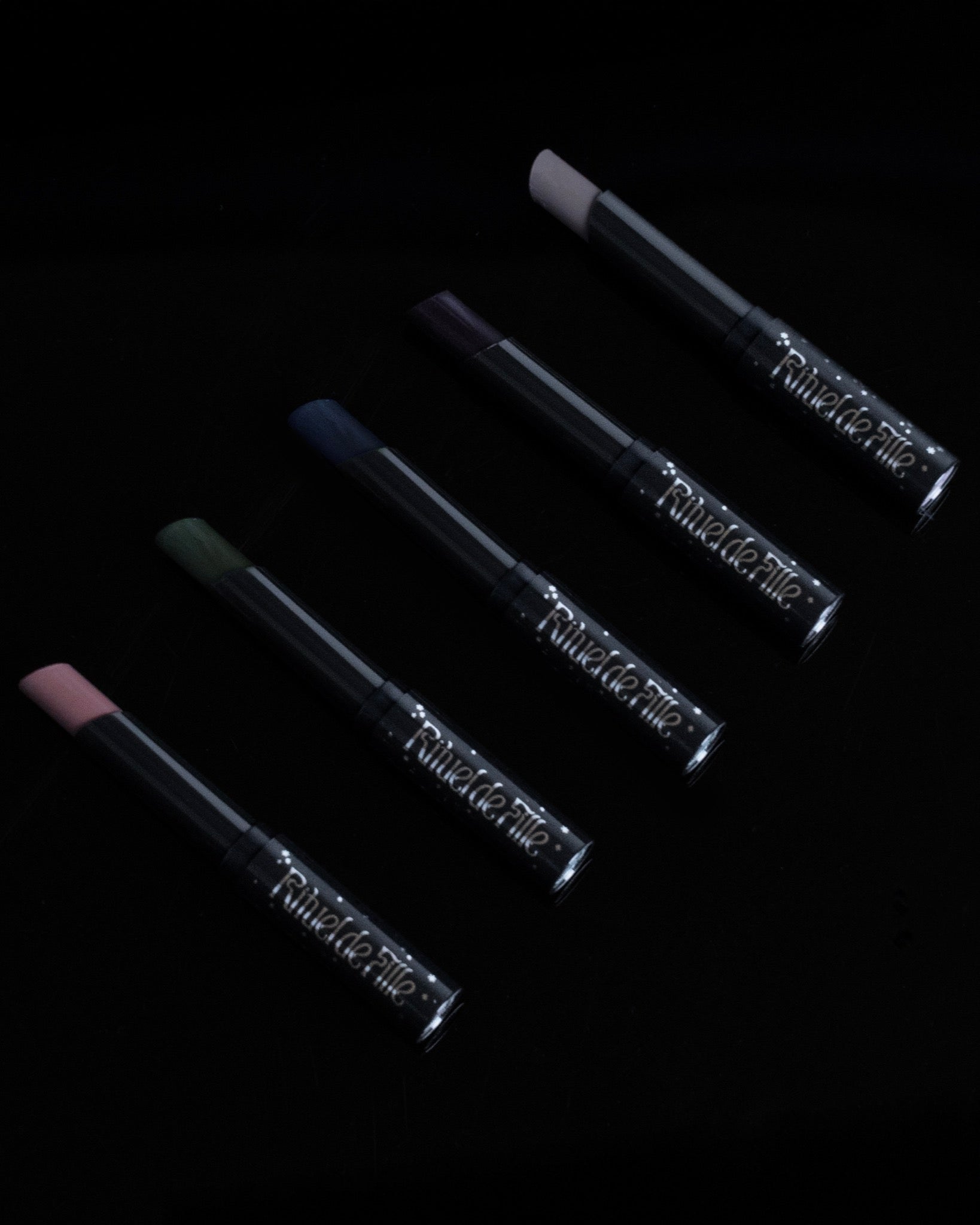 Four slim cosmetic tubes arranged diagonally on a black background, each with a different color tip visible: pink, green, blue, and dark purple. The tubes are metallic silver with embossed text that appears to read 'F.